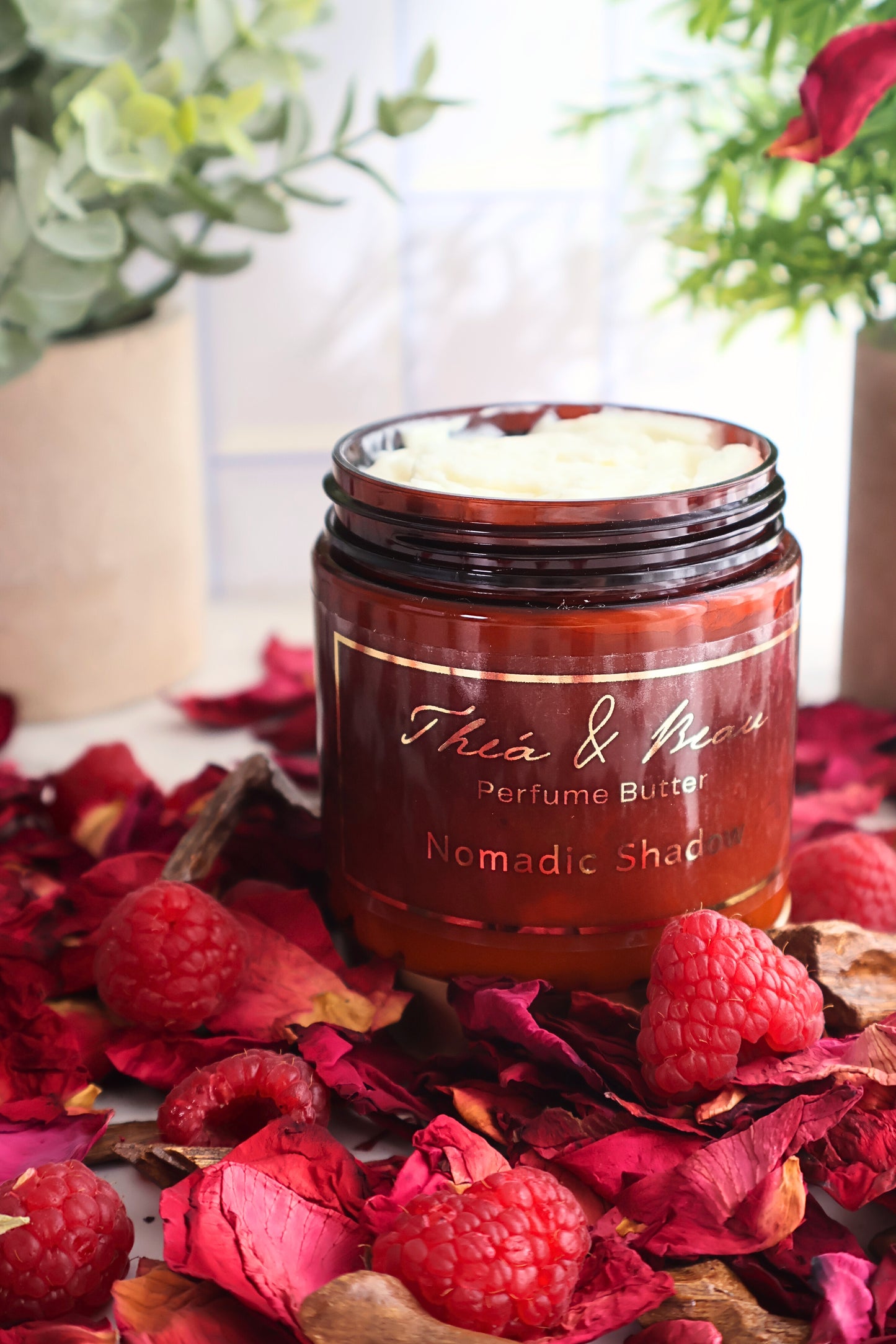 Close-up of Nomadic Shadow perfume body butter showing back of jar on white surface with raspberries rose petals and oud accents