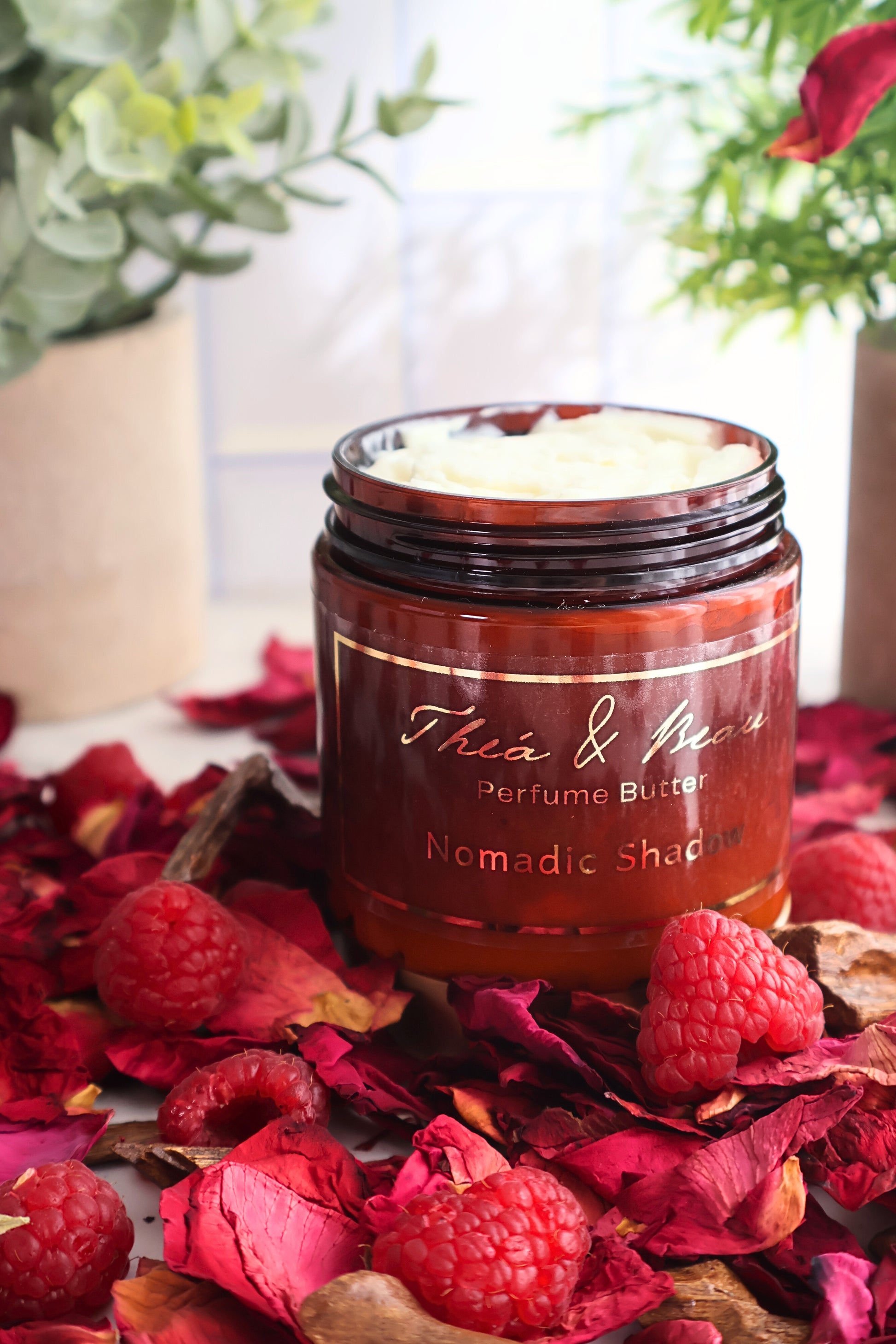 Close-up of Nomadic Shadow perfume body butter showing back of jar on white surface with raspberries rose petals and oud accents